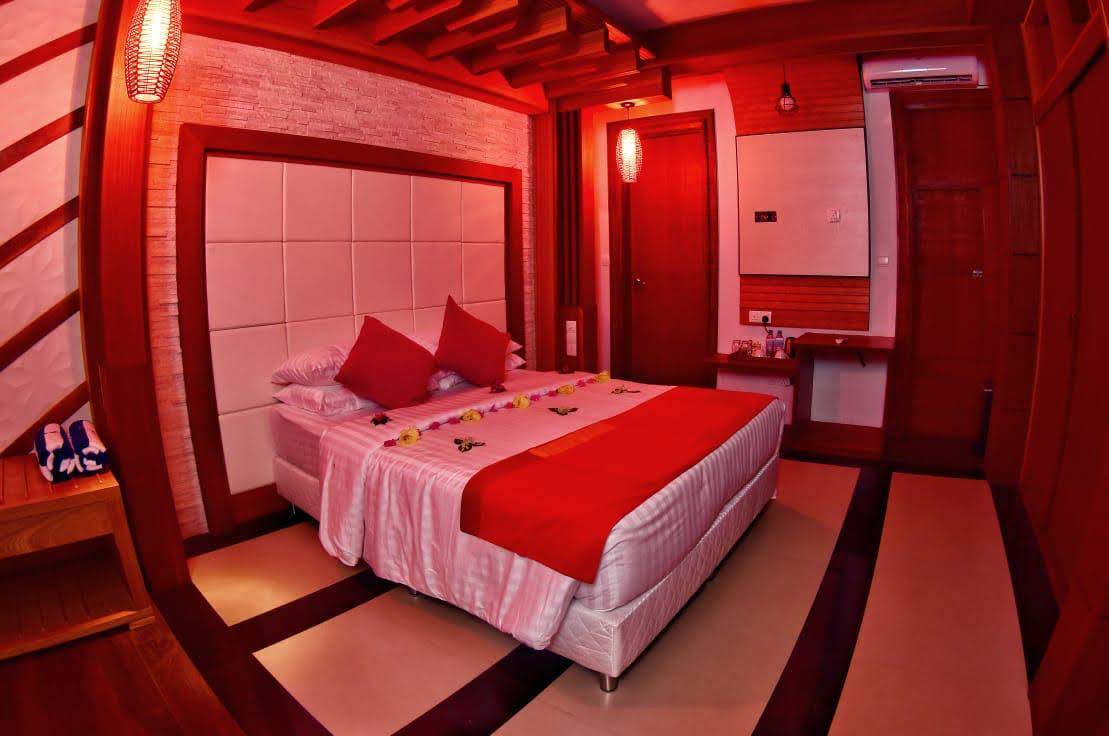 A well-appointed bedroom features a spacious bed covered with soft linens and accent pillows. Red lighting creates a warm ambiance, while decorative elements like flowers are arranged on the bed. Wooden accents and a minimalist design enhance the room's contemporary aesthetic.