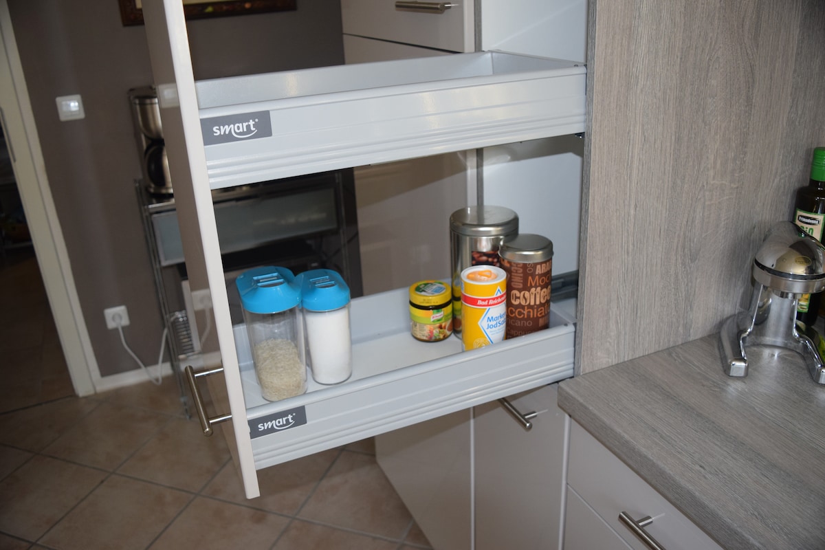 A modern kitchen storage unit features two pull-out shelves, showcasing a variety of pantry items. Containers of sugar, flour, and coffee are neatly organized alongside canned goods, all set within a sleek cabinet against a textured wall.