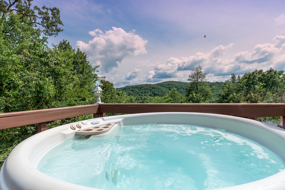 The hot tub is positioned on the deck, offering views of the surrounding mountains. The water appears inviting and is surrounded by a wooden railing. Lush greenery is visible in the background against a sky filled with soft clouds.