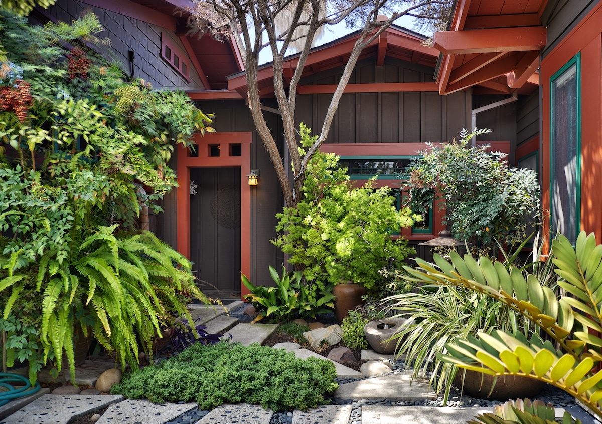 A serene courtyard features a variety of lush greenery, including ferns and flowering plants. A stone pathway leads to a private entrance, framed by vibrant red and dark wood architecture. Natural elements blend harmoniously with artistic details, creating a tranquil outdoor space.