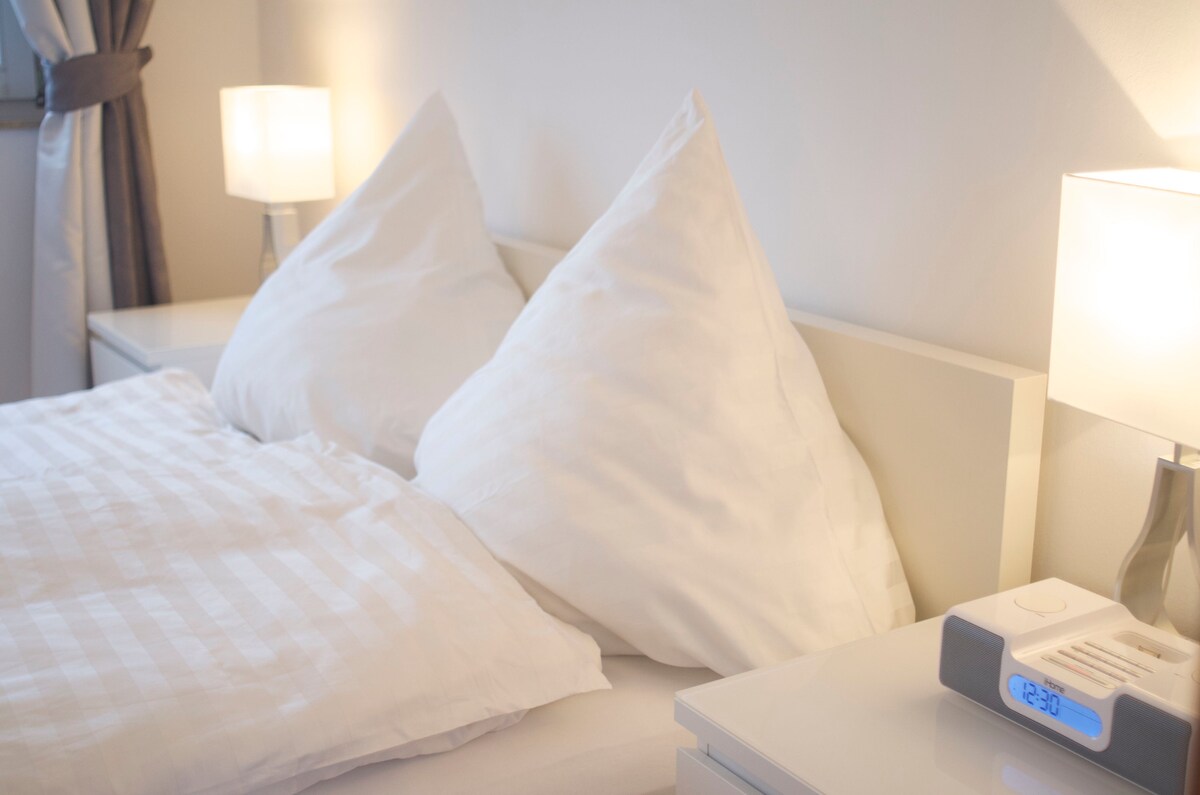 A spacious bed is dressed in light, crisp linens with two large, unique pillows prominently displayed. Soft bedside lamps provide gentle illumination, enhancing the calming atmosphere of the cozy sleeping area.