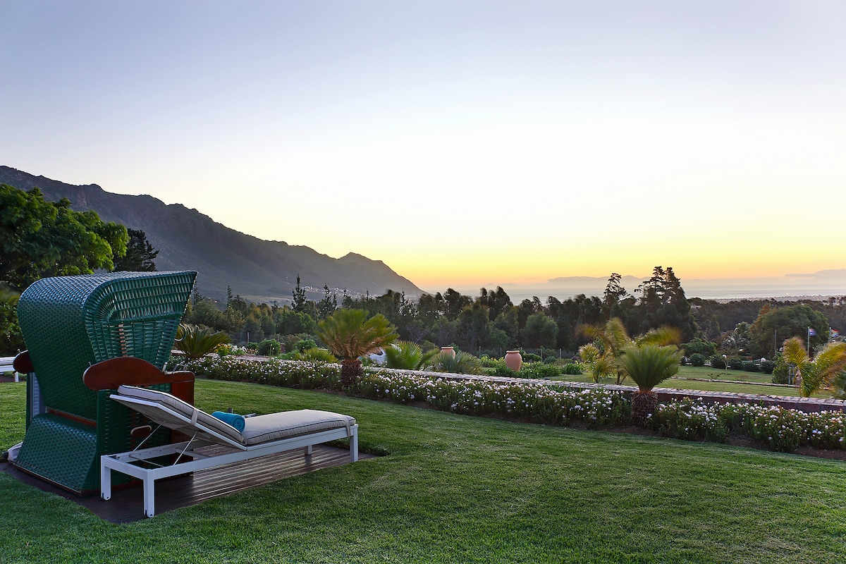 A lounge chair is positioned on a grassy area, overlooking a scenic landscape that includes rolling hills and distant mountains. The sky transitions from warm hues to cooler tones as the sun sets, providing a serene backdrop to the lush green gardens and palm trees.