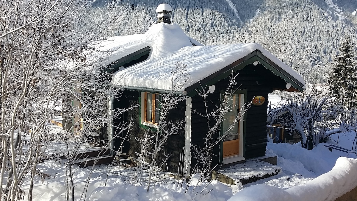 The chalet is nestled amidst a snowy landscape, featuring a charming exterior with a sloped roof and wooden accents. Snow covers the ground and branches, while large windows provide a glimpse of the warm interior. Scenic mountains rise in the background, enhancing the peaceful atmosphere.