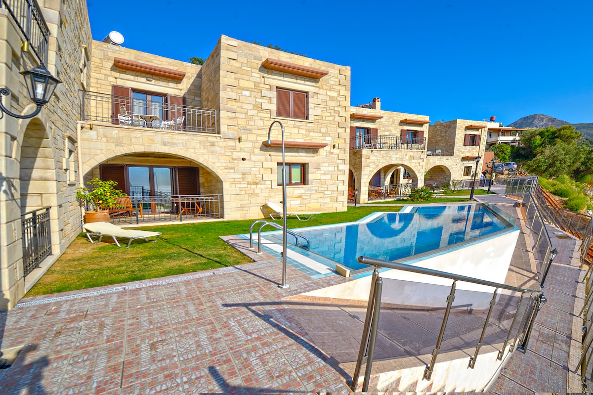 An outdoor pool is positioned among stone villas, surrounded by green lawns and paved walkways. The villas feature large windows and balconies, hinting at inviting outdoor spaces. Sun loungers are arranged beside the pool, ensuring a place for relaxation in the warm sun.