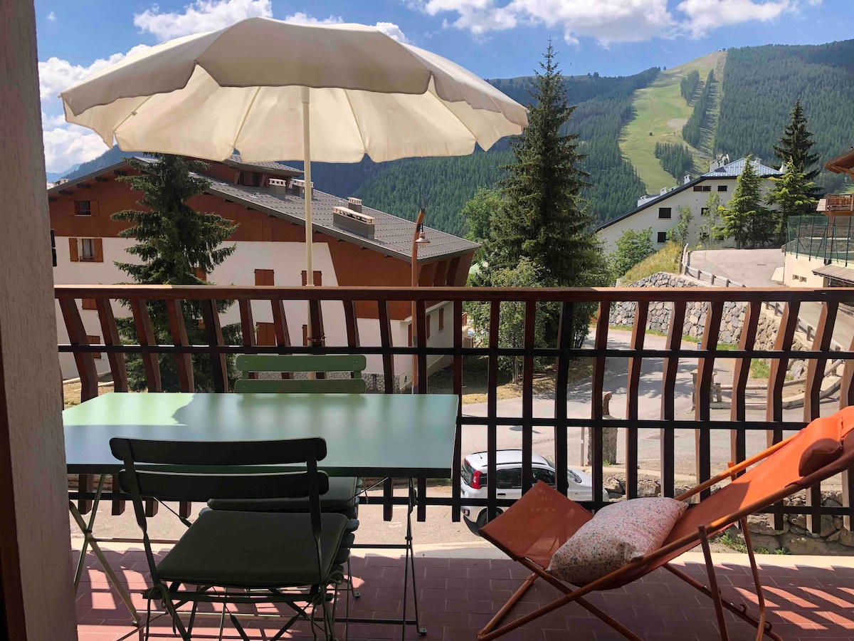 A private terrace is furnished with a dining table and folding chairs, overlooking mountain scenery. An umbrella provides shade, and a lounge chair adds a comfortable seating option. Surrounding greenery and distant ski slopes contribute to a peaceful outdoor atmosphere.