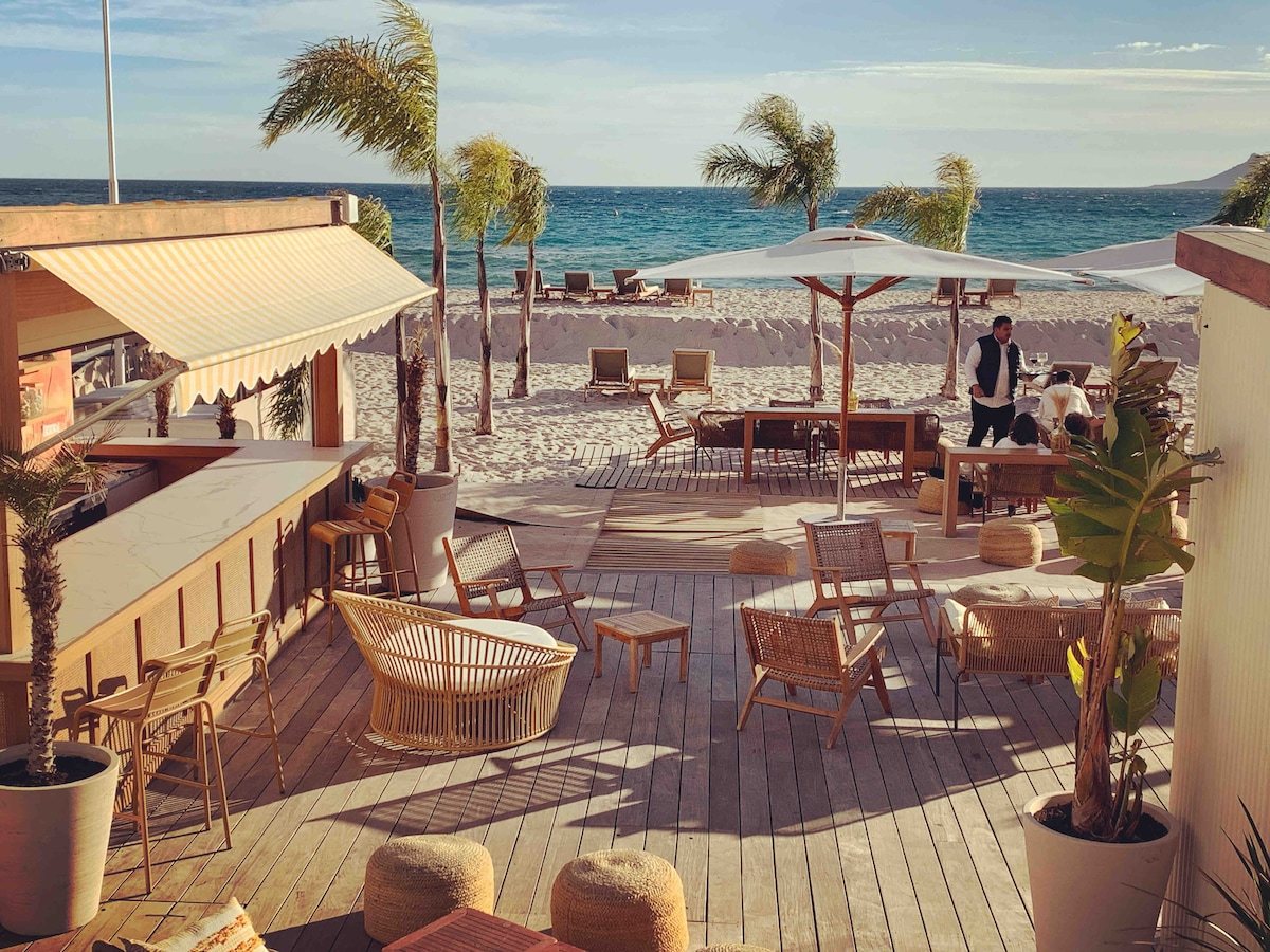 An outdoor beach lounge area is showcased, featuring wooden decking and comfortable seating arrangements. Sun loungers and umbrellas are placed on the sandy beach, with palm trees providing ambiance. A bar area is visible, enhancing the inviting space for relaxation by the ocean.