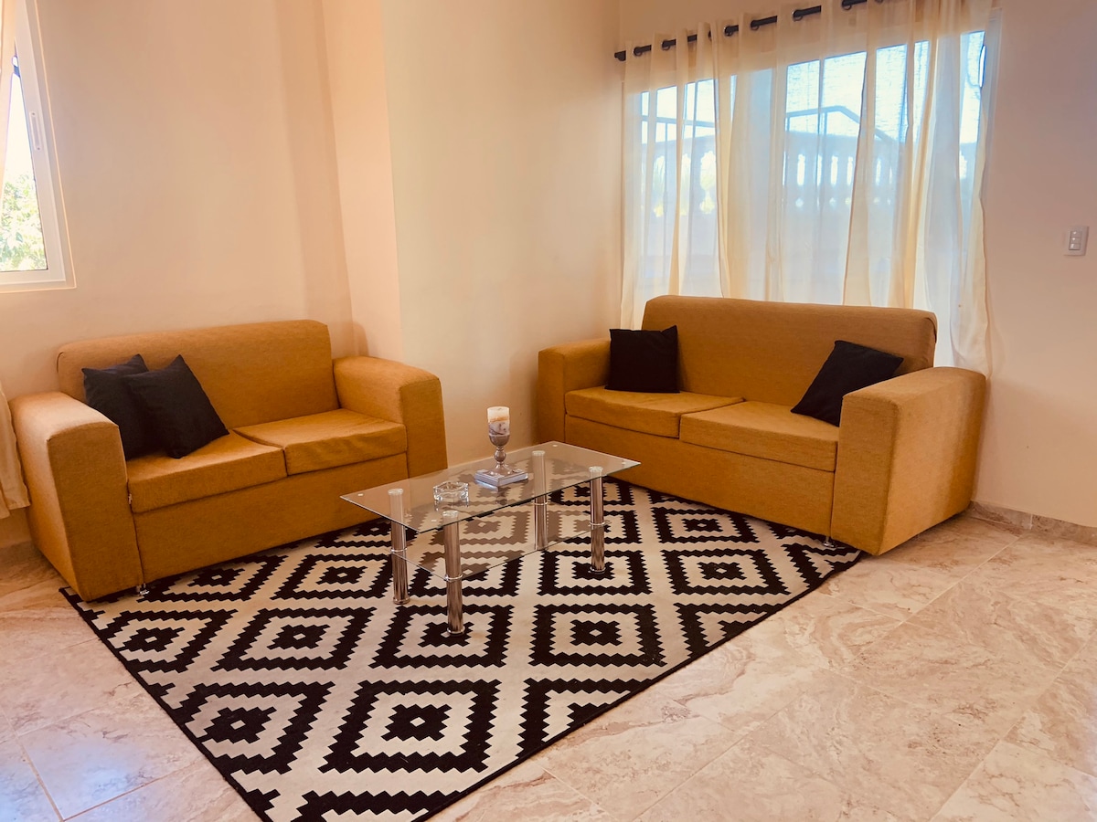 A cozy seating area is presented with two brown sofas, accented by black cushions. A glass coffee table sits at the center, topped with decorative items. A patterned black and white rug covers the floor, while natural light filters through sheer white curtains.