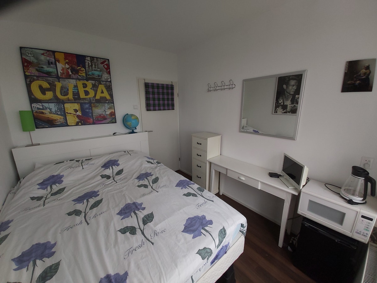 A bright bedroom is equipped with a spacious bed featuring floral bedding. A mirror and a simple desk are positioned adjacent to a microwave and kettle. Artwork titled 'CUBA' adorns the wall, complemented by a globe and practical storage options.