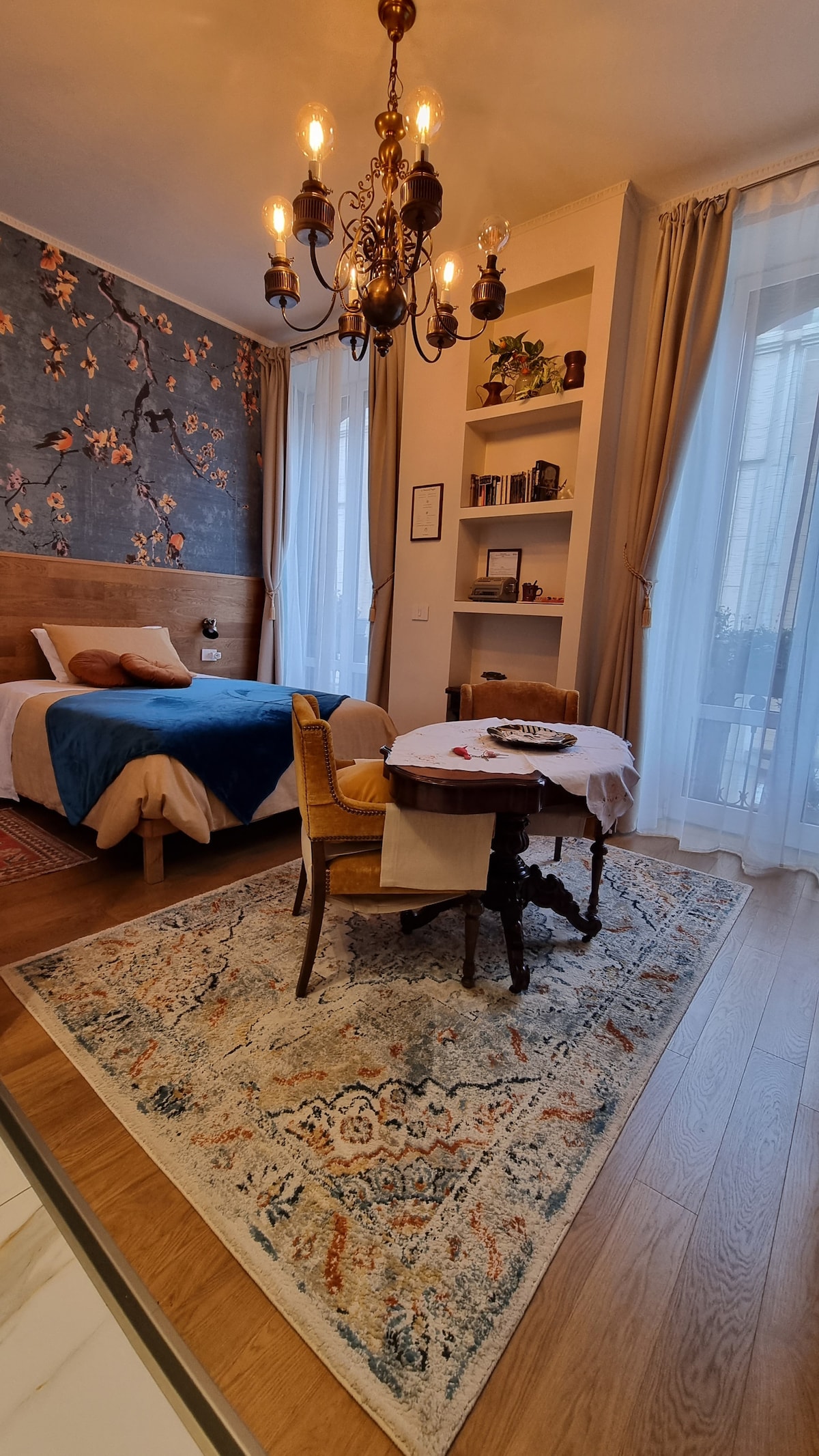 A cozy room features a spacious bed with neutral bedding, complemented by a blue throw. A chandelier hangs from the ceiling, illuminating the floral wallpaper. A wooden table with chairs offers a dining space, while large windows allow natural light to fill the room.