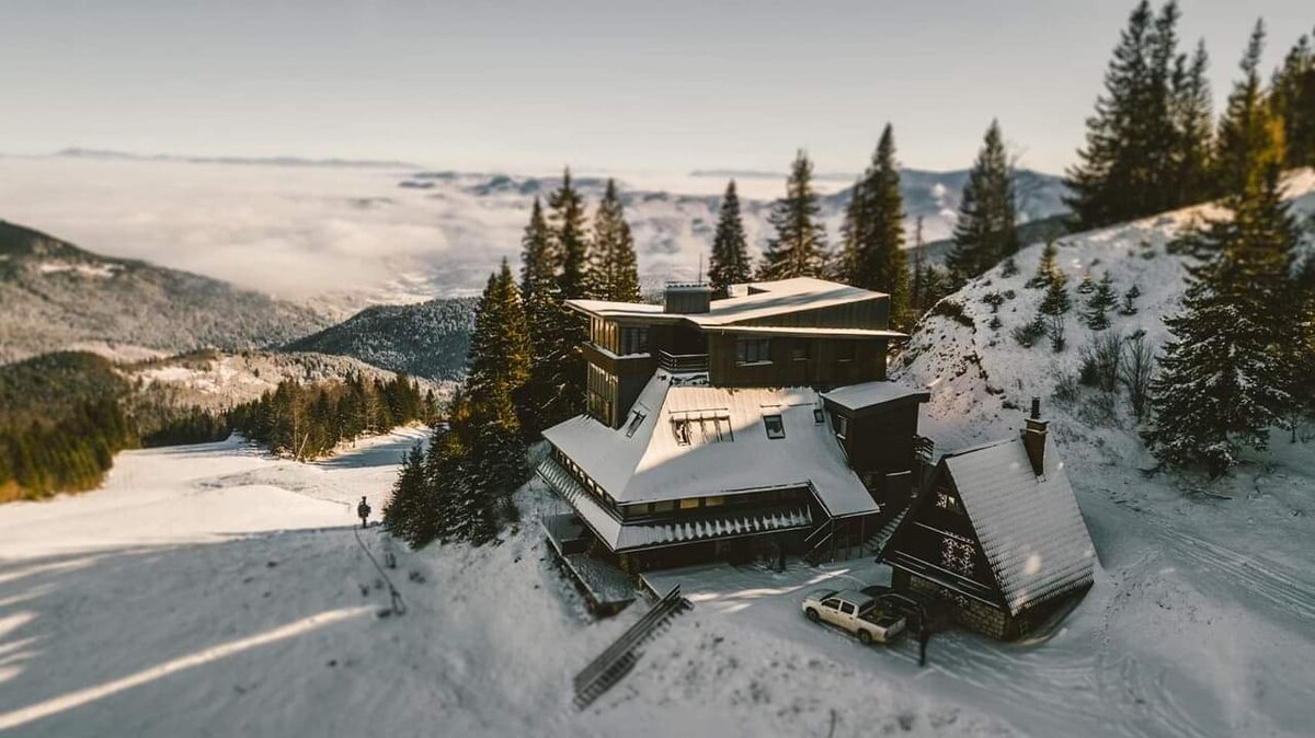 An aerial view captures the cozy apartment nestled in a snowy landscape, surrounded by tall trees. The two distinct building sections feature sloped roofs, with a serene ski slope visible in the foreground, creating a blend of natural beauty and winter charm.