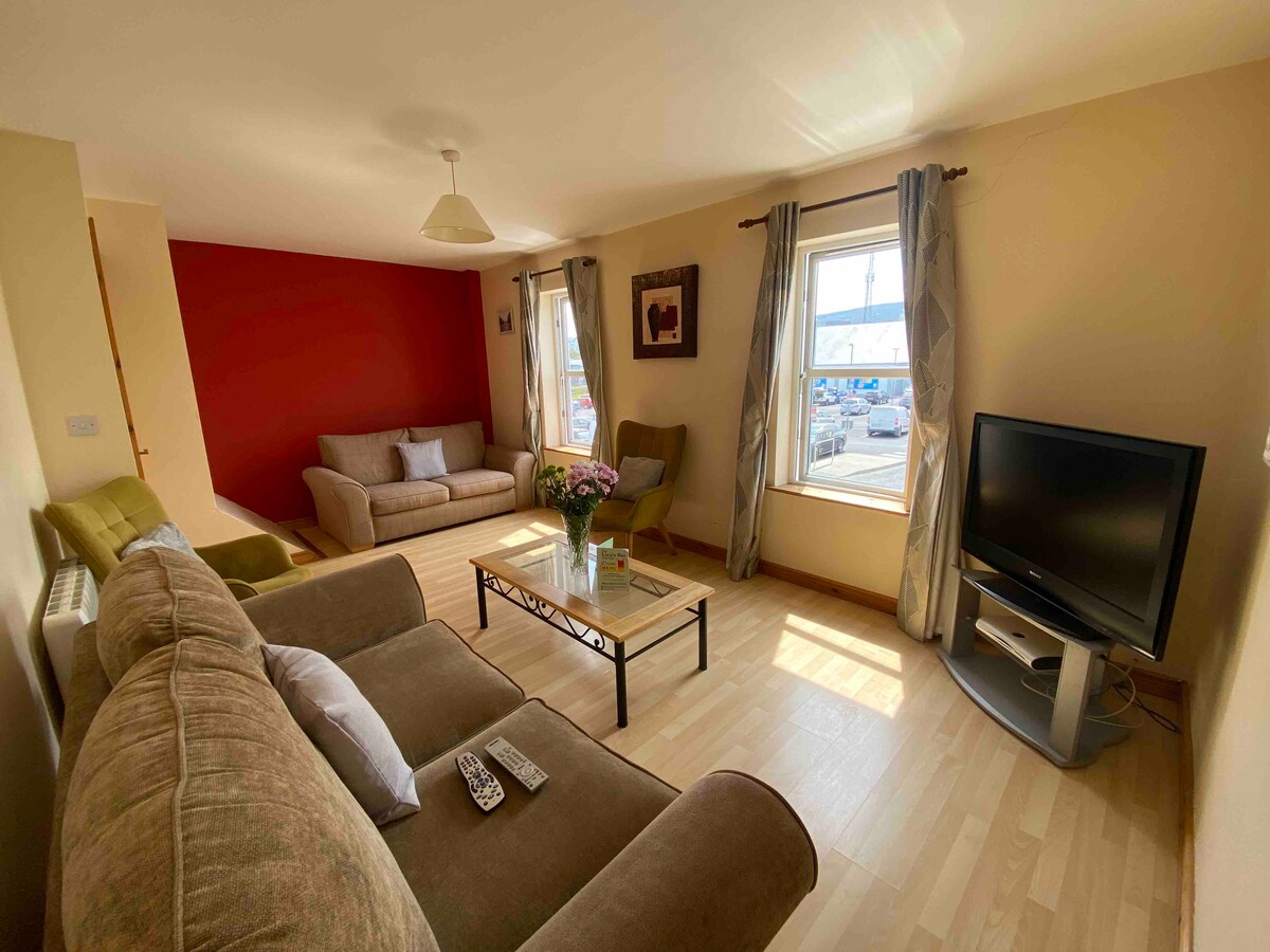 The living room features a mix of comfortable seating including a spacious couch and armchairs. Natural light enters through the large windows, illuminating the wooden flooring. A coffee table sits centrally with a vase of flowers, complemented by a television mounted nearby.