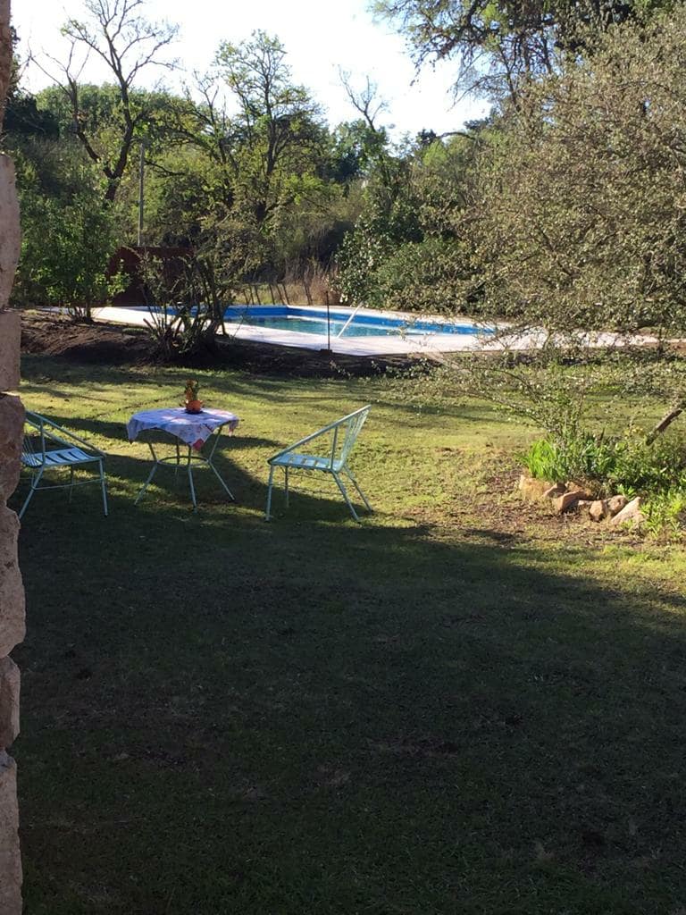 A serene outdoor space is highlighted featuring scattered greenery and a swimming pool in the background. A small table with chairs is positioned on the grass, providing a peaceful spot for relaxation amidst nature.