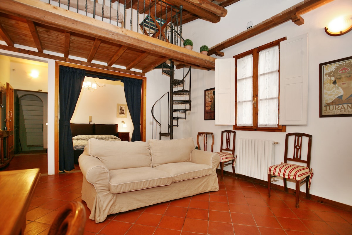 A spacious living area is highlighted by a cream-colored sofa, with a staircase leading to a wooden loft above. Natural light enters through a window adorned with white curtains. The room features a dining table and four chairs positioned nearby.