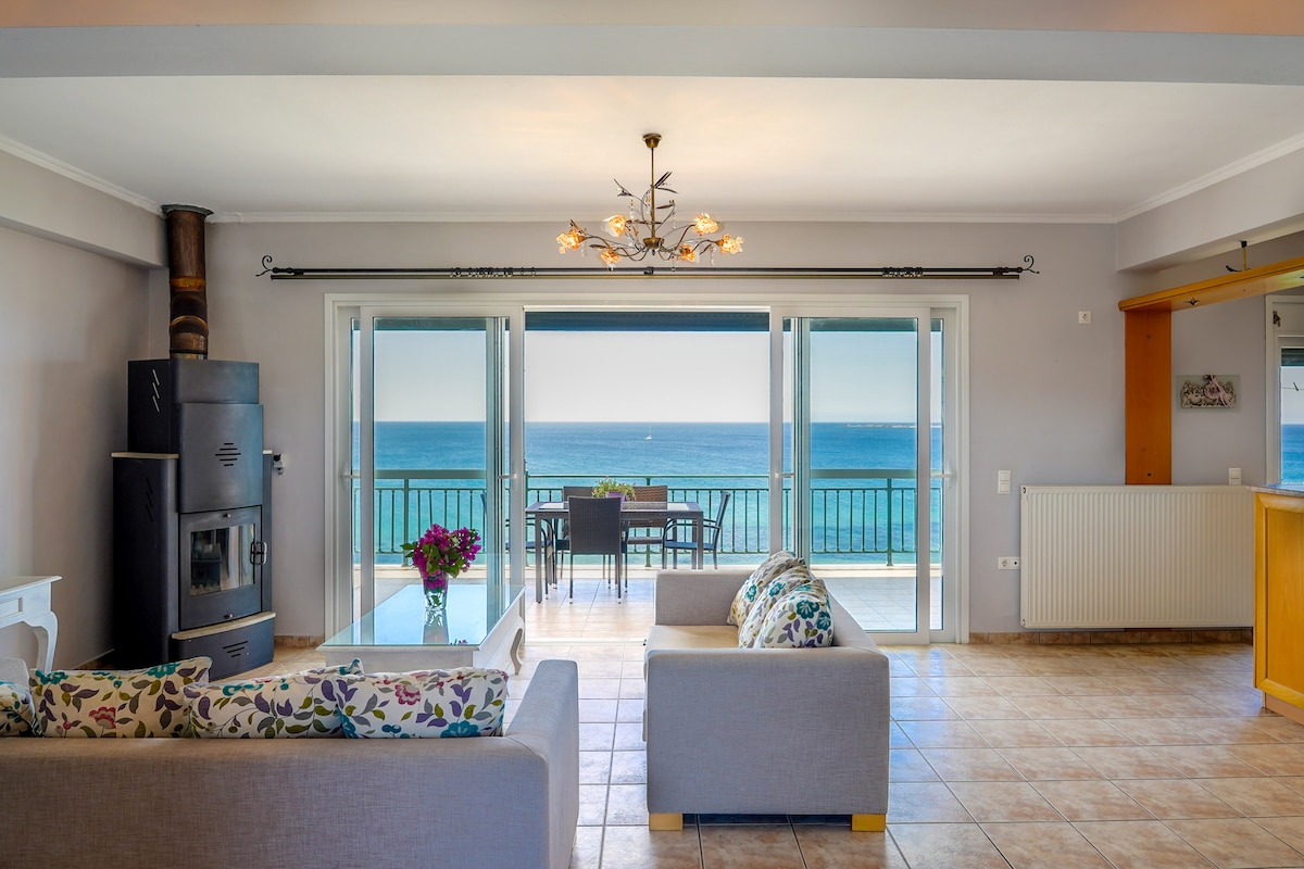 A spacious living area features large glass doors that open to a balcony with stunning sea views. Comfortable seating is arranged around a coffee table, and a decorative light fixture adds visual interest. The warm color palette enhances the inviting atmosphere.