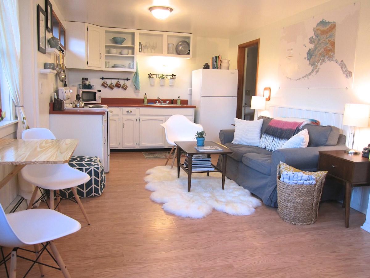A cozy living area features a gray couch adorned with colorful throw pillows. A small coffee table sits atop a soft, fluffy rug. A dining table is accompanied by two chairs, and kitchen appliances are visible in the background, providing convenient access to cooking amenities.