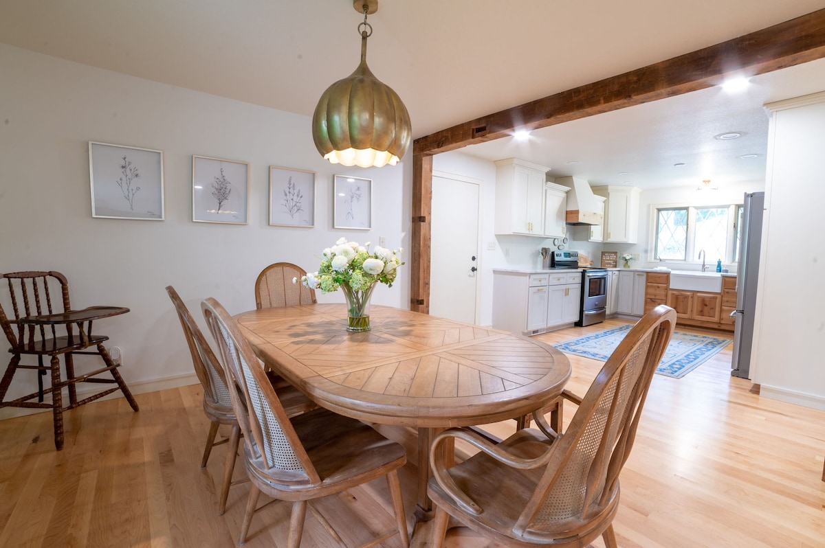 A dining area features a large, round wooden table surrounded by six woven chairs. A vase of flowers sits at the center, while a decorative light fixture hangs overhead. The open layout leads to a bright kitchen with white cabinetry and stainless steel appliances.