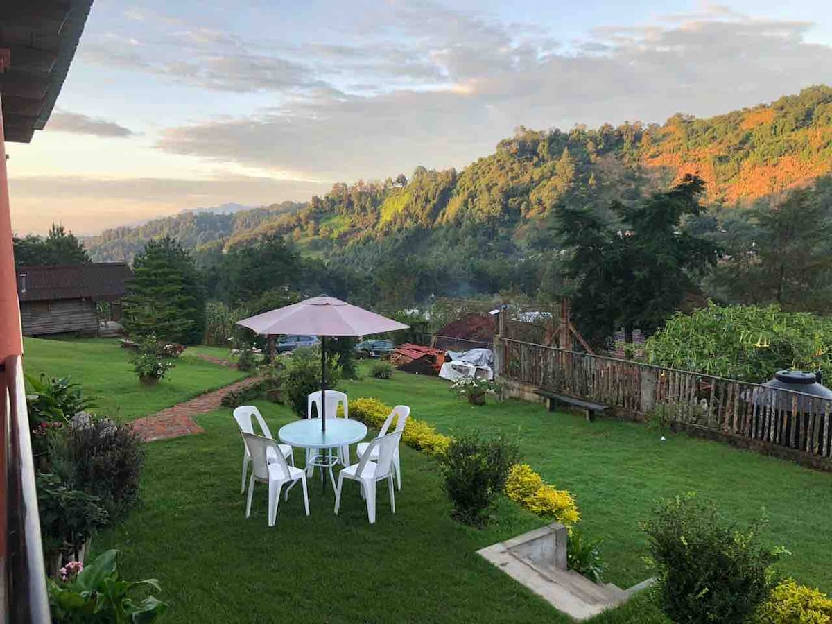 An outdoor garden area features a round table with four white chairs beneath a pale umbrella. Lush green grass surrounds the seating arrangement, and distant mountains are visible in the background, complemented by a colorful landscape of trees and foliage.