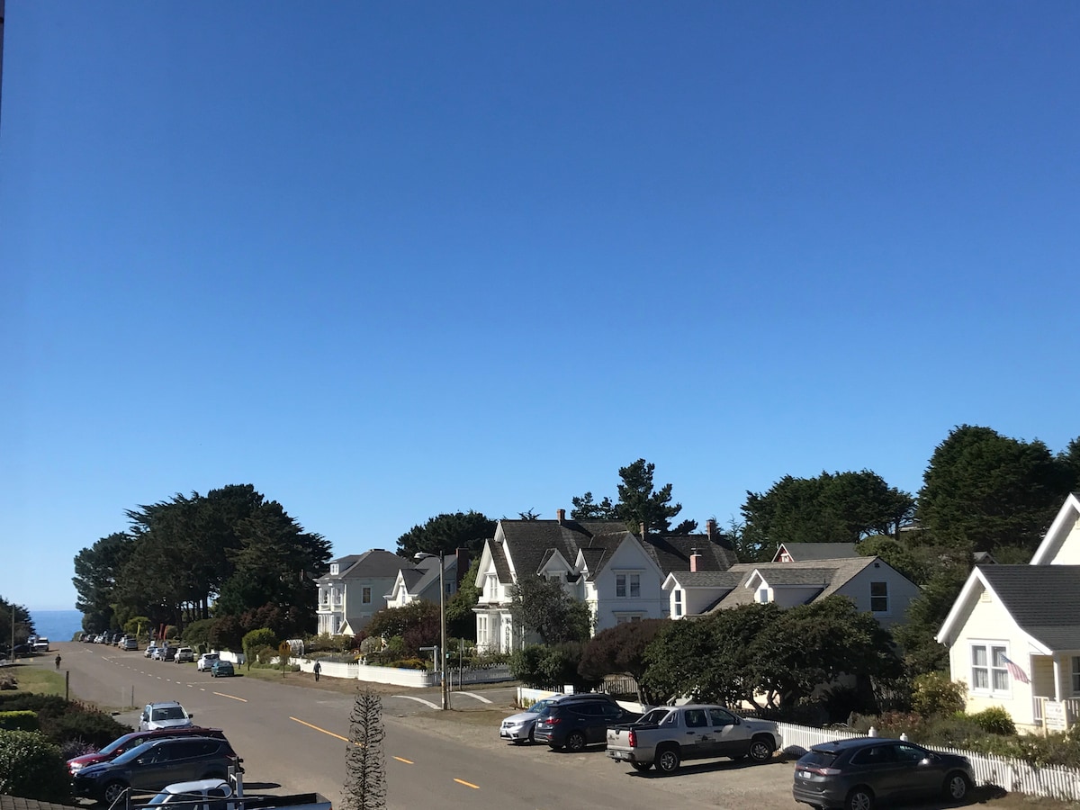 The image captures a clear blue sky above a tree-lined street featuring well-maintained homes with varied architectural styles. Parked vehicles line the street, and lush greenery offers a scenic backdrop, contributing to the welcoming feel of the neighborhood.