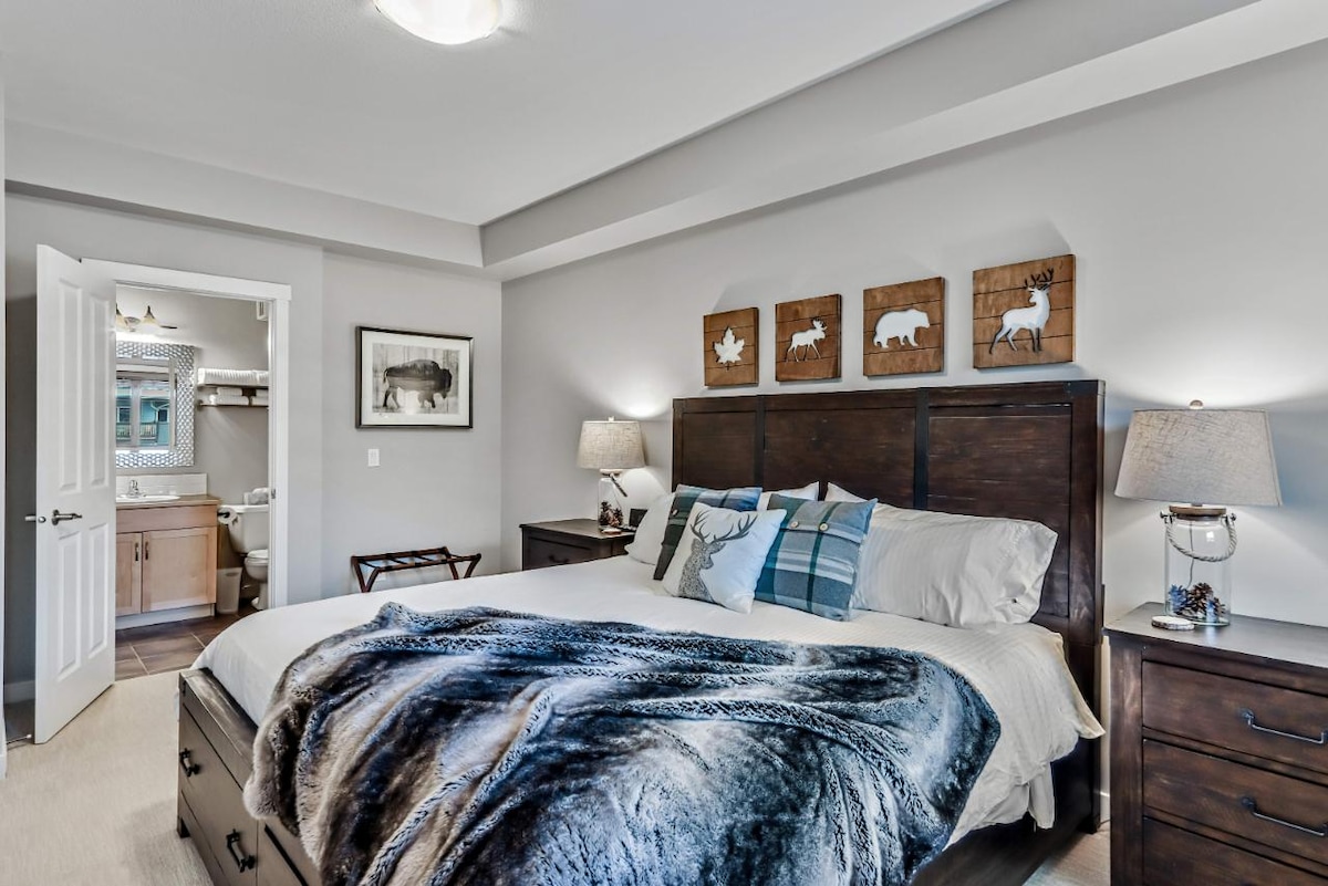 A serene bedroom is highlighted by a king-sized bed adorned with layered blankets and decorative pillows. Rustic wooden art decorates the wall above the headboard. Nightstands with soft lighting flank the bed, while a doorway leads to a bathroom area, enhancing the overall comfort.