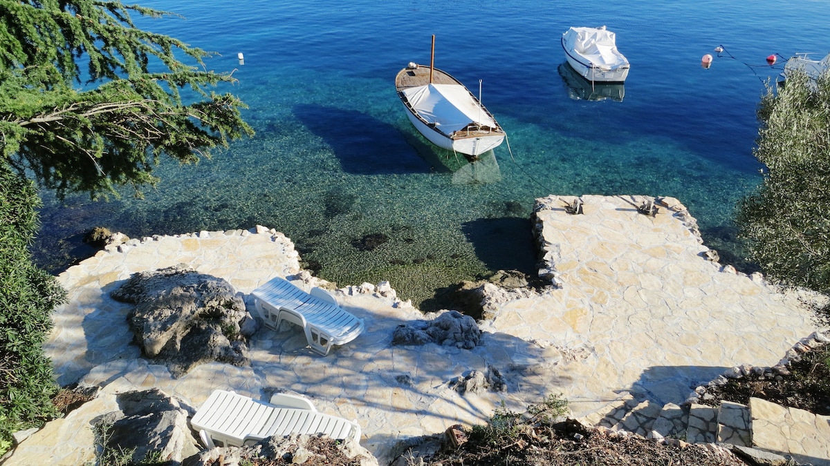 A serene waterfront area features a rocky shore with laid stone pathways. Two boats are moored nearby in clear blue water. Loungers are positioned on the sunlit terrace, inviting relaxation by the sea.