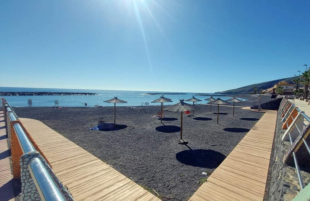 The image depicts a serene beachfront scene with several thatched umbrellas dotting the dark, sandy shore. Wooden pathways provide access along the water's edge, leading to calm ocean views under a clear blue sky.