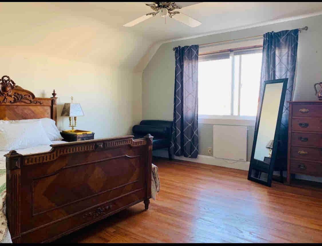 A spacious bedroom features a vintage wooden bed with intricate carvings and a decorative blanket. A full-length mirror stands against the wall, next to a sleek black chair. Light enters through a large window, framed by patterned curtains, creating a bright and airy atmosphere.