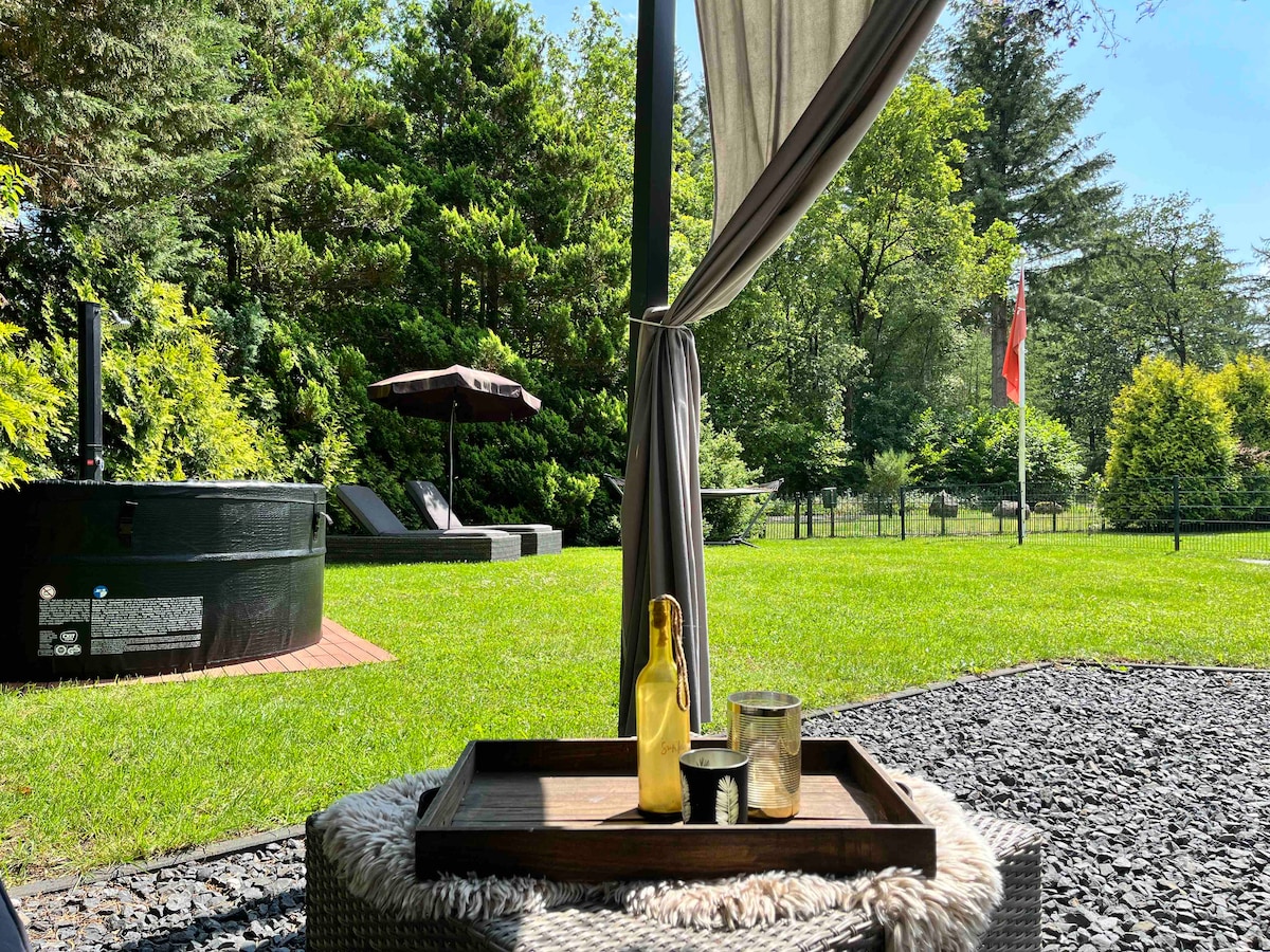 A serene outdoor space features a jacuzzi surrounded by a lush green lawn. A shaded lounge area is visible with a sun umbrella and comfortable reclining chairs. A small table holds a bottle and glasses, providing a perfect spot for relaxation amidst nature.