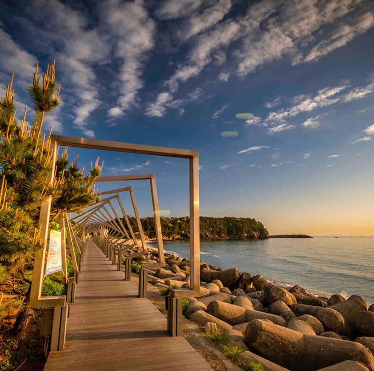 A wooden walkway extends along the coastline, framed by modern metal structures. The pathway is flanked by smooth boulders and greenery, leading towards a tranquil sea. The sky is filled with soft clouds, reflecting warm colors from the sunset.