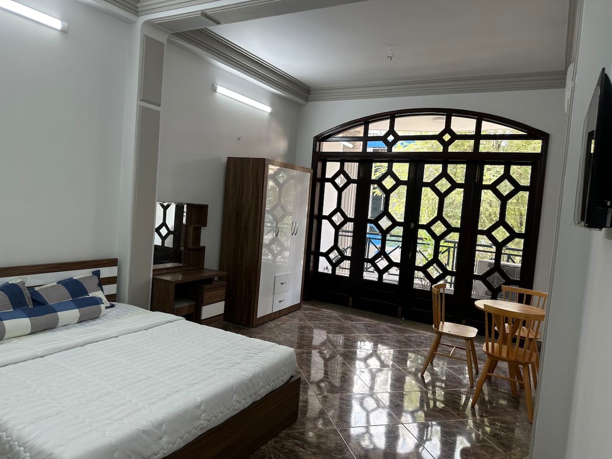 The room features a spacious bed with a blue and white checkered throw blanket. A wooden table and two chairs occupy one corner. Large windows with decorative panes allow natural light to fill the space, illuminating the shiny tiled floor.