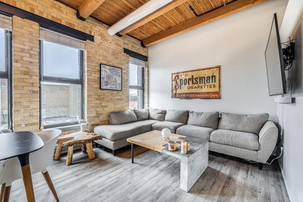 An inviting living area features a plush sectional sofa and a wooden coffee table. Exposed brick walls complement the ceiling beams, while large windows allow natural light to fill the space. A wall-mounted television enhances the modern decor, and a dining table is visible in the background.
