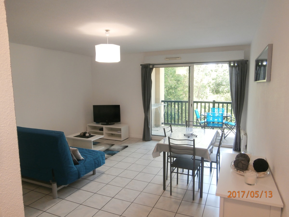 A spacious living area is displayed, featuring a convertible sofa and a flat-screen television. A dining table with seating is arranged near large glass doors that open to a balcony, which overlooks a tree-lined view. Neutral tiled flooring complements the light walls.
