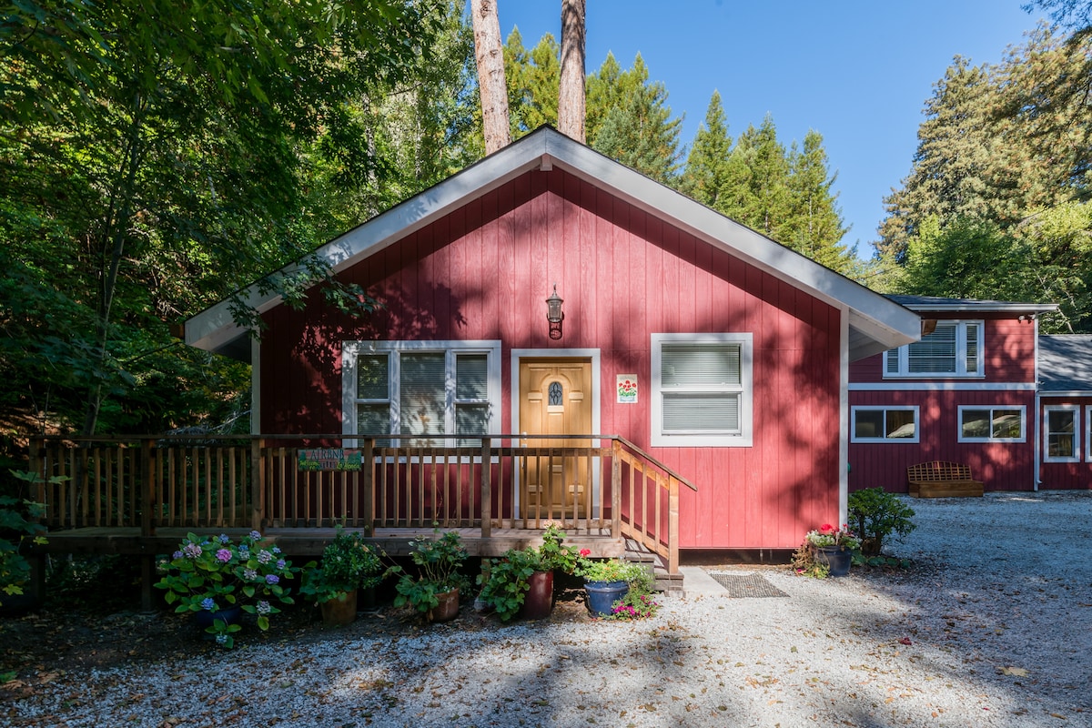 The exterior of a red cottage is showcased, surrounded by lush greenery. A wooden deck with a sturdy railing leads to the front door, which is centered beneath a soft light fixture. Colorful flowers in pots are arranged at the base of the porch.