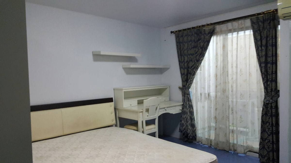 A light blue room features a spacious bed without bedding and a simple wooden desk with a chair. Sheer curtains cover the window, allowing natural light to enter. Floating shelves are mounted on one wall, adding a touch of functionality.