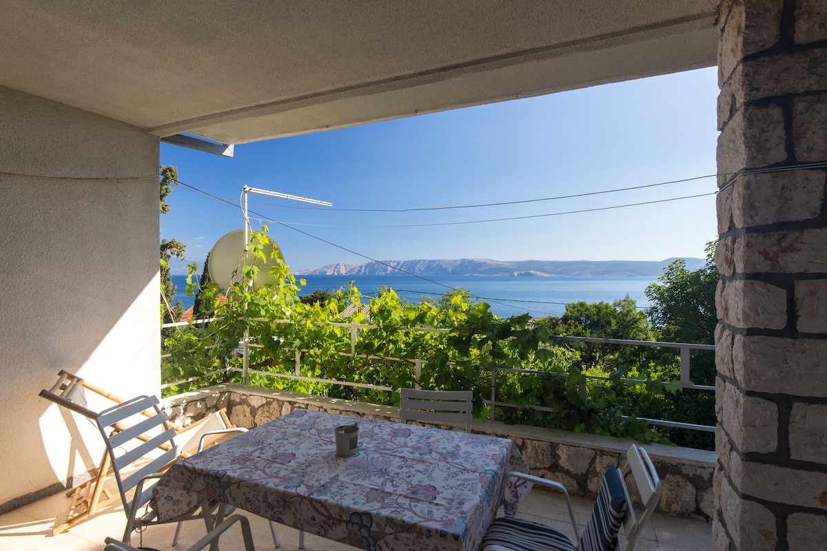A spacious terrace is framed by greenery and offers views of the sea and distant mountains. A table is set beneath a floral tablecloth, accompanied by several chairs. Sunlight filters through the leaves, creating a bright and welcoming atmosphere.