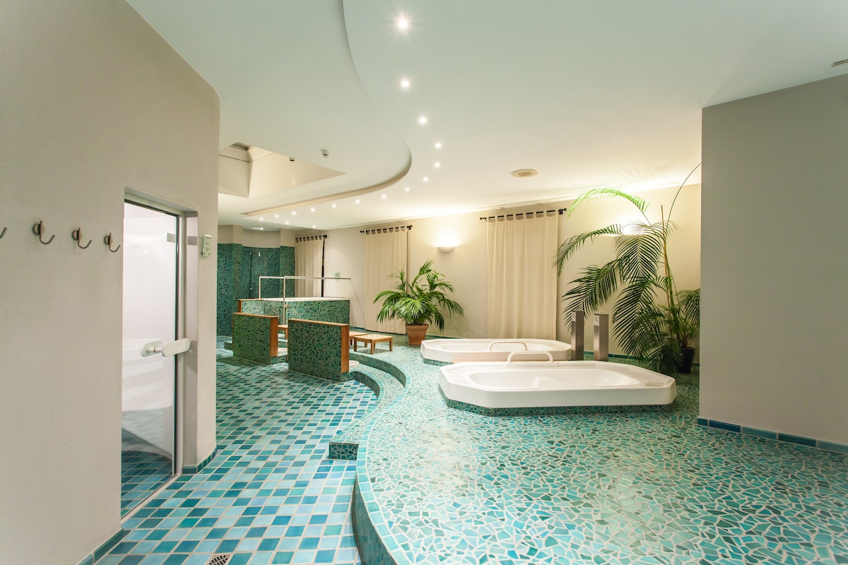 A spacious wellness area is featured, with elegant blue mosaic tiles covering the floor. Two whirlpool tubs are positioned nearby, surrounded by potted plants. Soft lighting creates a serene atmosphere, while a steam room is accessible through a glass door.