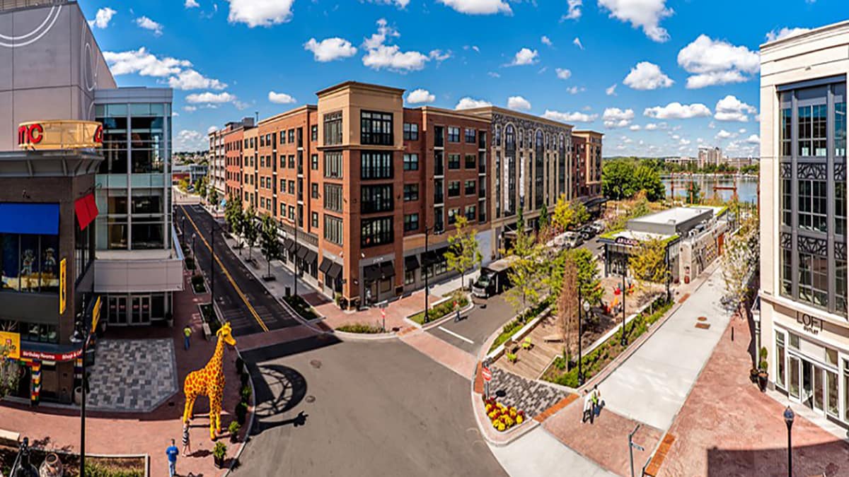 A vibrant urban scene is depicted, featuring a well-maintained street lined with modern buildings. Outdoor seating areas are present along with landscaped greenery. A large giraffe sculpture is positioned prominently, contributing to the lively atmosphere. Bright blue skies and fluffy white clouds complete the view.