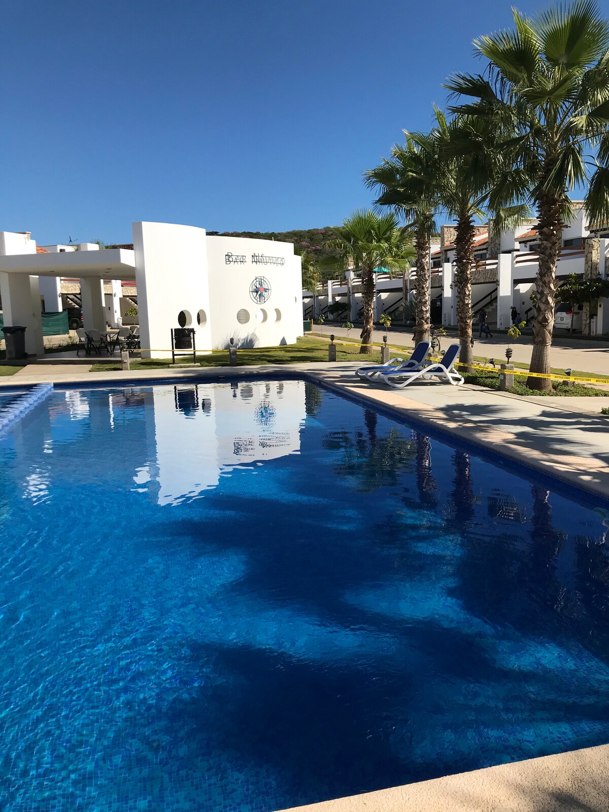 A tranquil swimming pool reflects the clear blue sky, surrounded by well-maintained landscaping and palm trees. Lounge chairs are positioned along the poolside, inviting relaxation. The clean lines of the property’s architecture are visible in the background, enhancing the serene atmosphere.