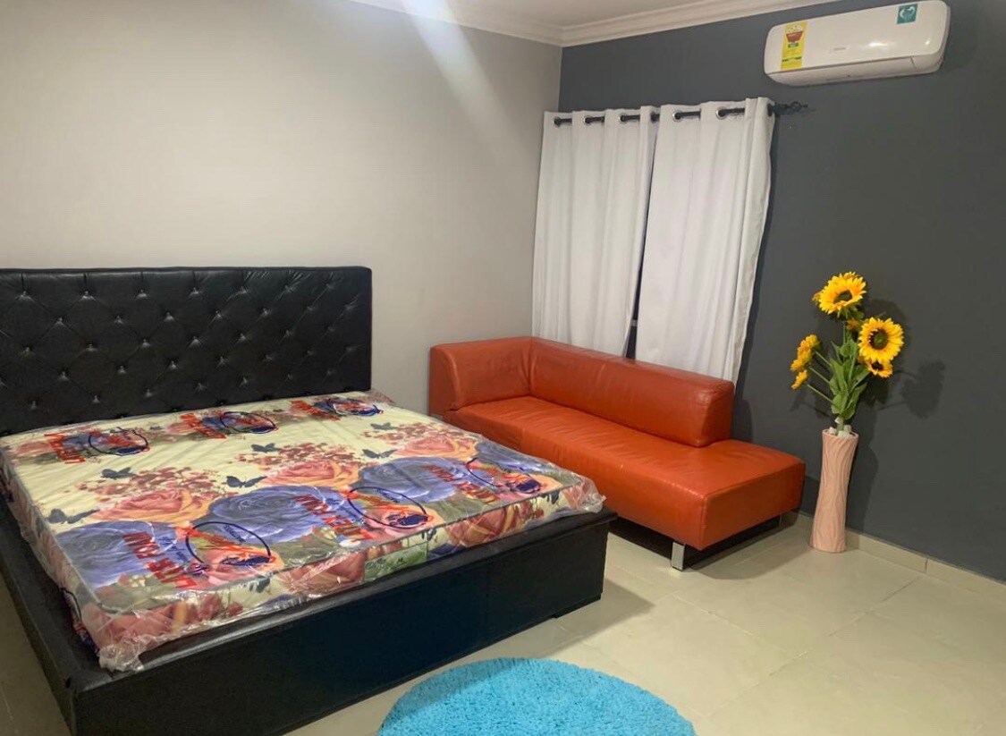 A spacious sleeping area features a large black bed adorned with a colorful patterned mattress. An orange sofa sits against the wall, accompanied by a vase displaying bright sunflowers. Curtains frame a window, and a round blue rug lies on the tiled floor.