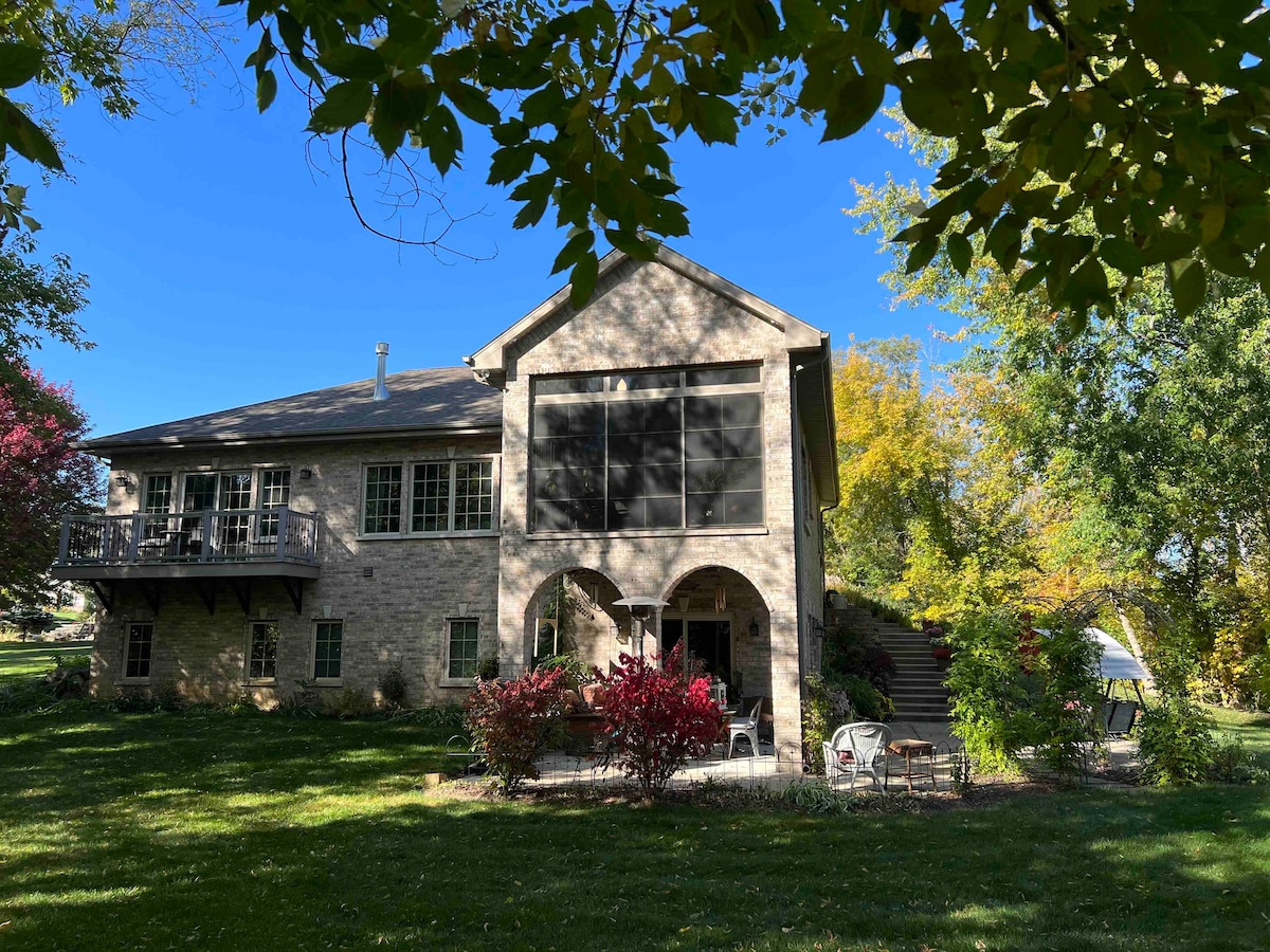 The exterior of the home features a two-story brick structure with large screened windows and arched entrances. Lush greenery surrounds the property, complemented by vibrant fall foliage. A spacious patio area with seating is visible, set against a backdrop of mature trees and a well-maintained lawn.