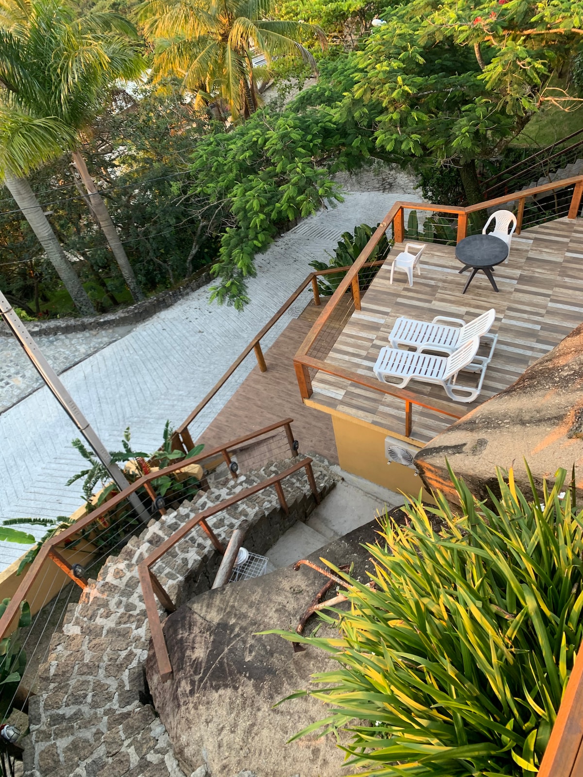 An elevated view reveals a spacious deck with two white lounge chairs and a small table, surrounded by lush greenery and palm trees. A winding stone pathway leads to the entrance of the chalet, contributing to a tranquil outdoor setting.