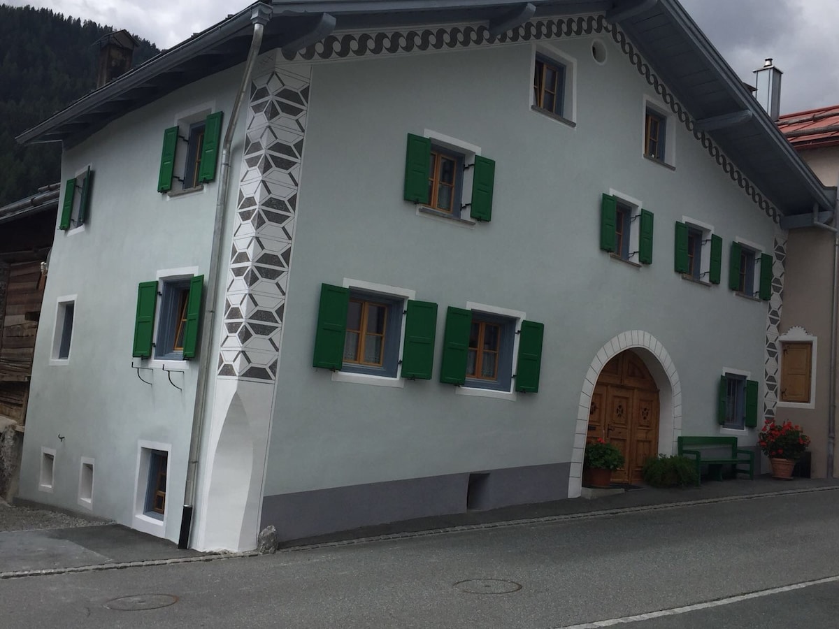 A traditional building is depicted with a charming façade featuring white walls and vibrant green shutters. Ornate architectural details complement the entrance, which is accentuated by a wooden door. The structure is situated along a quiet street, surrounded by picturesque mountain scenery.
