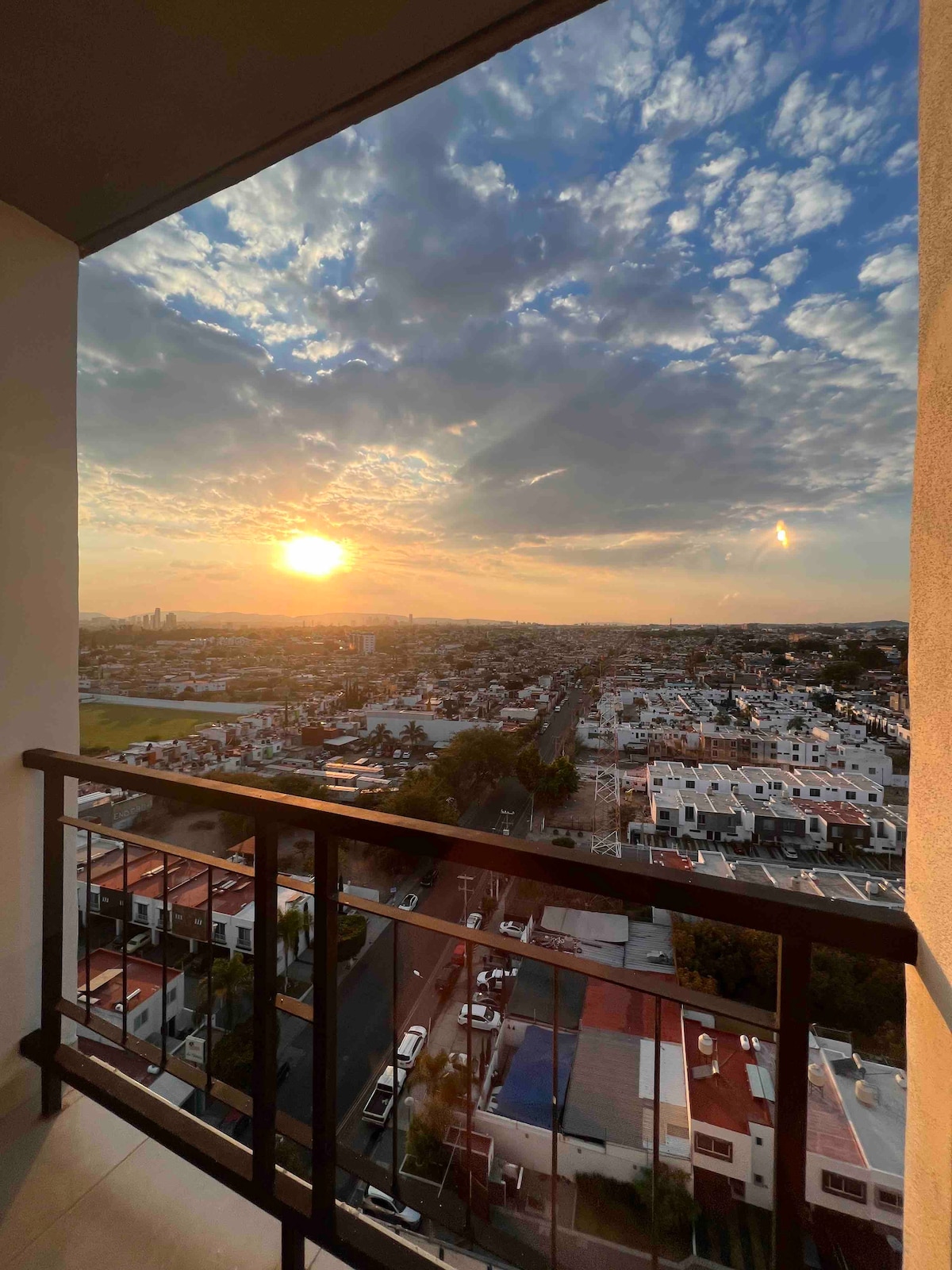 A balcony view reveals a sprawling cityscape during sunset, with warm hues illuminating the sky. A railing provides safety and an unobstructed perspective of rooftops and distant buildings. The scene captures a moment of transition from day to evening, emphasizing natural beauty.