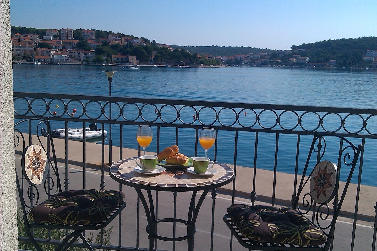A small balcony featuring a round table set with two cups and glasses of orange juice, alongside freshly baked pastries. Two decorative chairs with cushions provide comfortable seating, while a scenic view of the shimmering sea, distant hills, and charming village enhances the atmosphere.