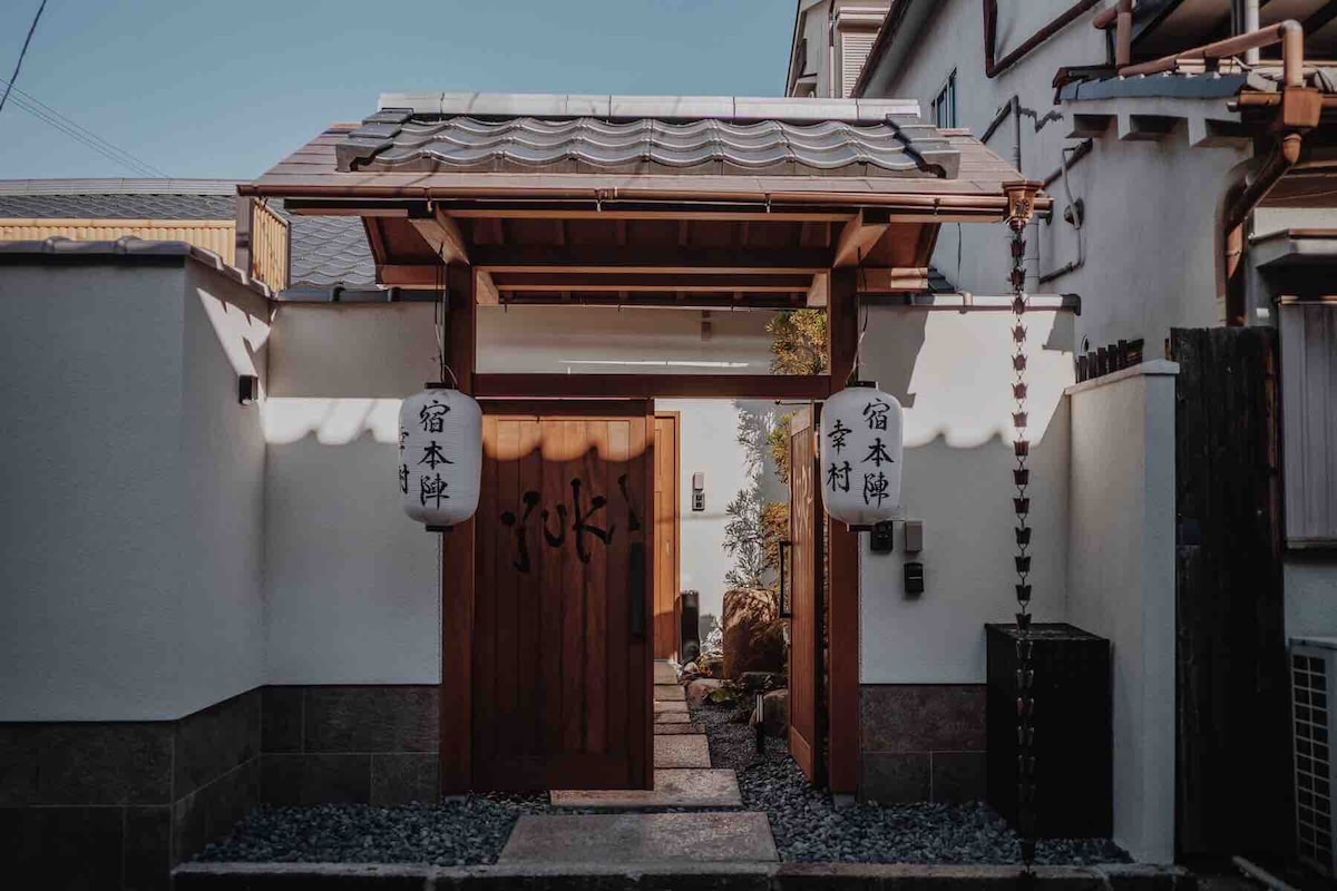 The entrance features a traditional wooden gate adorned with lanterns. A stone pathway leads through a serene garden, flanked by well-maintained greenery. The exterior reflects a blend of historical architecture and modern elements.