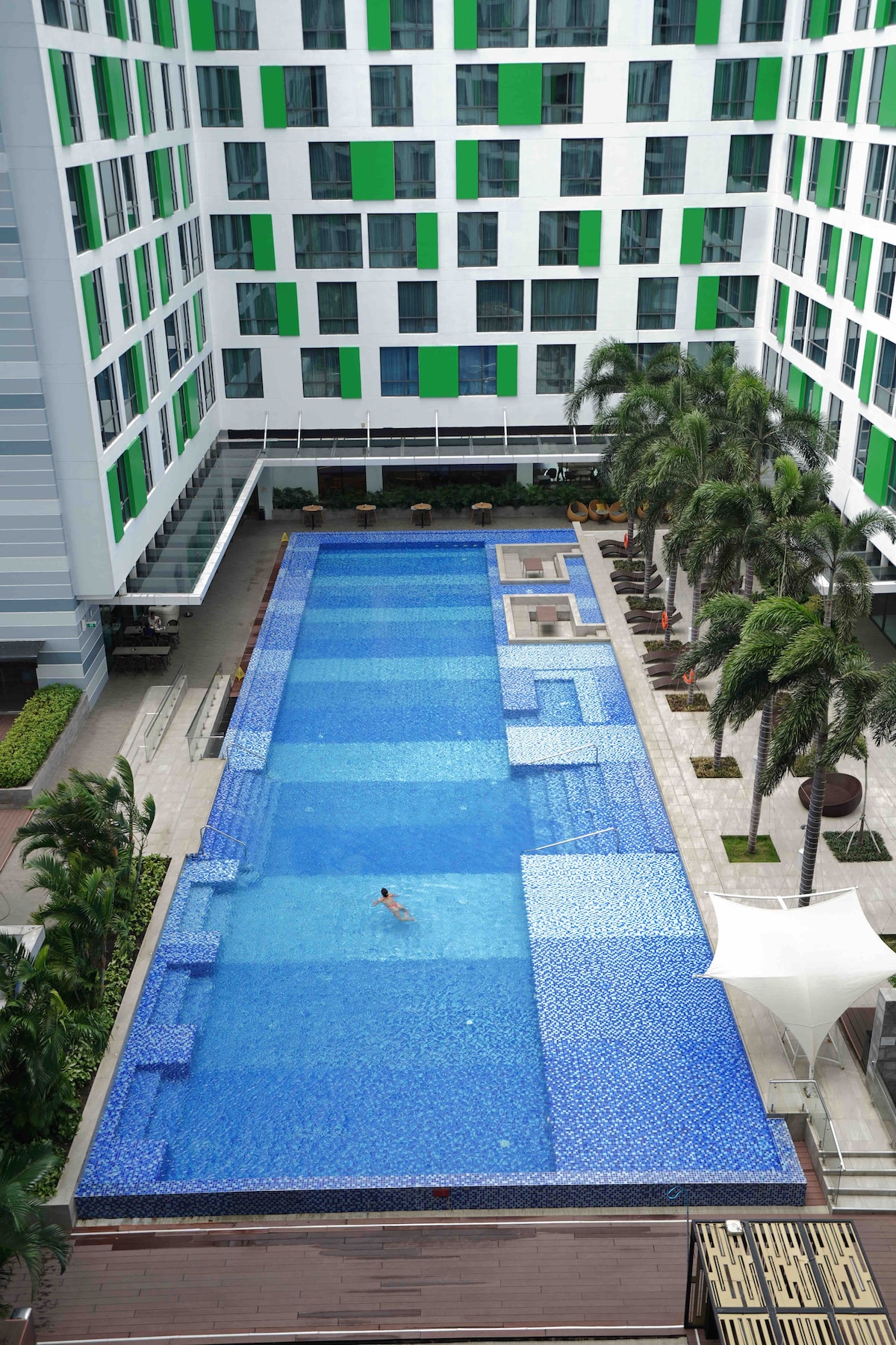 An outdoor swimming pool spans the courtyard, featuring varying depths and tiled sections. Sun loungers are placed along the pool's edges, surrounded by palm trees. A shaded area with seating is located at one end, providing a space for relaxation.