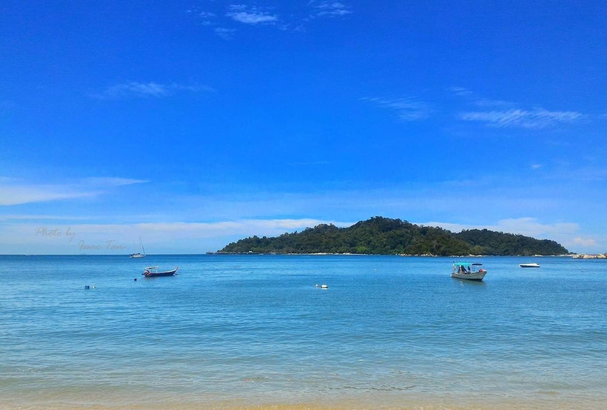 A serene seascape features calm turquoise waters under a bright blue sky. Small boats are anchored in the distance, with a lush, green island visible beyond the waterline, creating a peaceful coastal scene.
