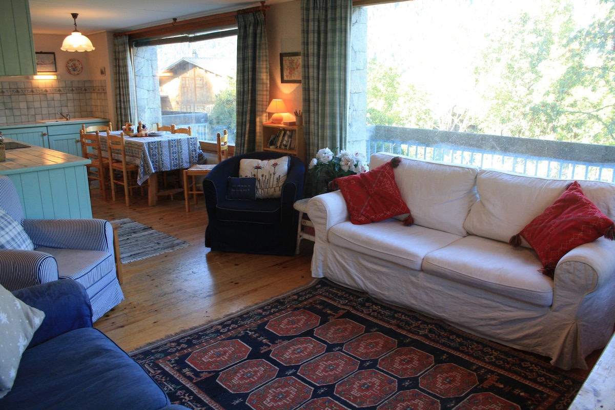 An open-plan living area features a cozy seating arrangement with a white sofa and dark blue armchair, adorned with red cushions. Large windows provide views of the surroundings, while a wooden dining table is set for six, complemented by bright curtains and wooden flooring.