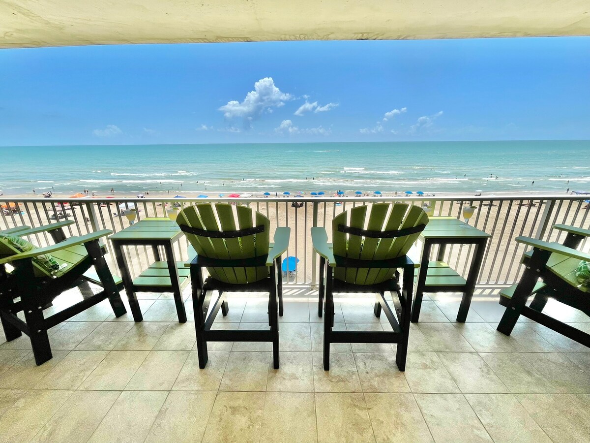 An expansive balcony offers a direct view of the Gulf of Mexico, framed by a railing. Four green adirondack chairs provide comfortable seating for guests to relax while enjoying the scenic beach landscape and ocean waves.