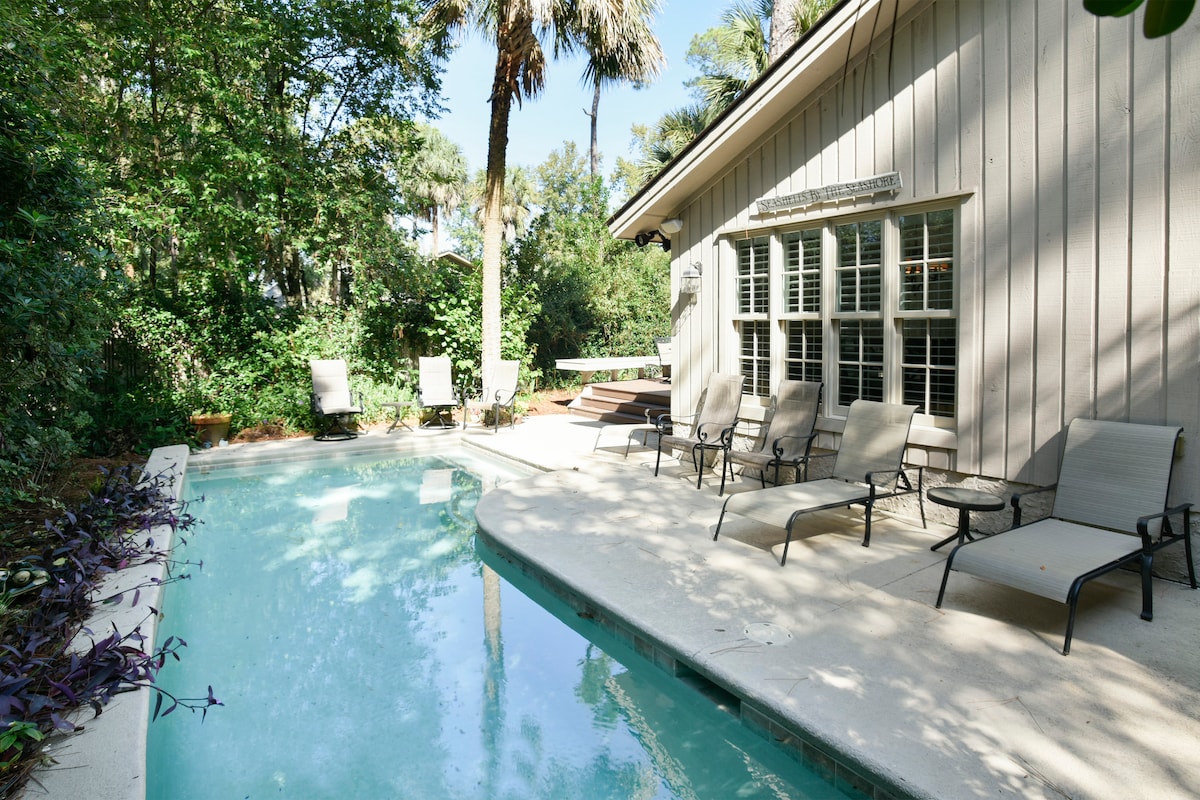 A private pool is surrounded by a lush landscape, with ample seating provided by several lounge chairs. Sunlight filters through the trees, creating a serene atmosphere near the exterior of the home, characterized by large windows and a spacious deck area.