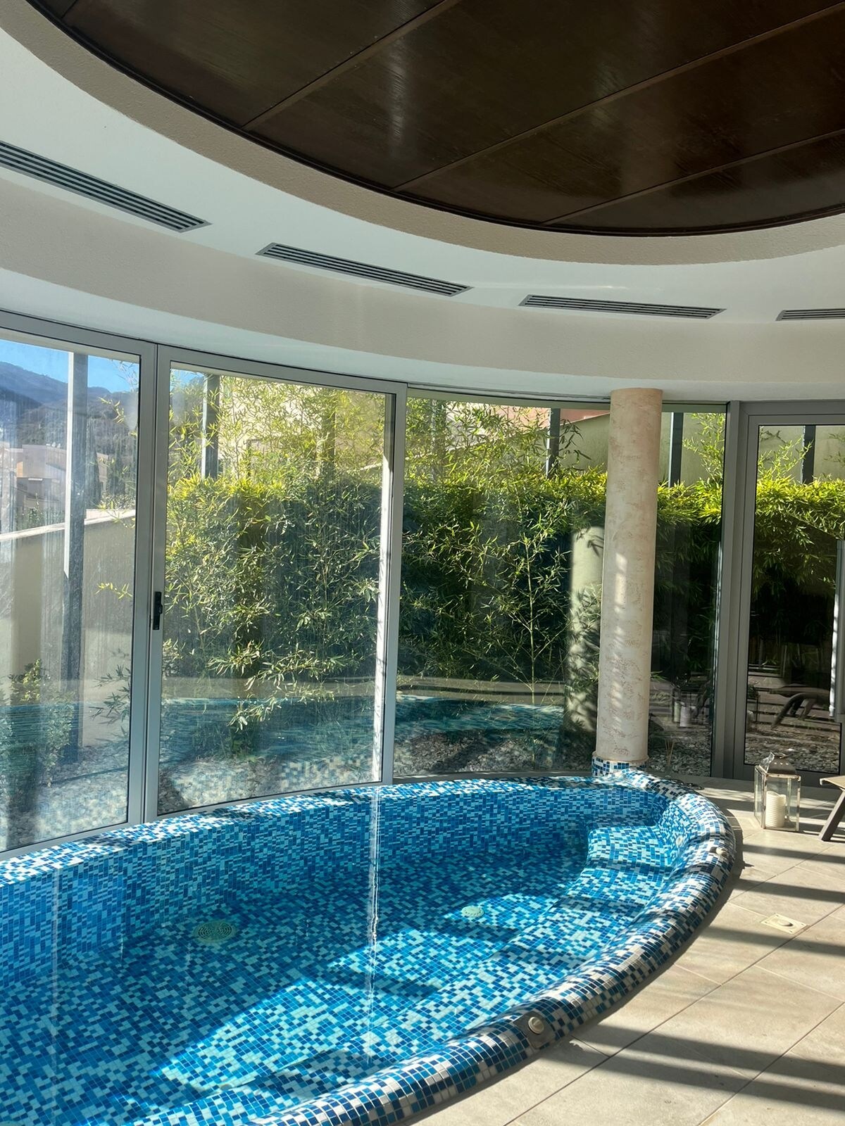 An indoor pool is featured, surrounded by large glass windows that offer views of lush greenery outside. The pool is adorned with blue mosaic tiles, and natural light floods the space, enhancing the serene atmosphere.